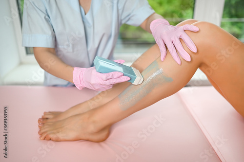 Woman with long tanned perfect legs and smooth skin having wax stripe depilation hair removal procedure on legs in beauty salon. Beautician in blue robe, pink gloves. Body care, epilation spa concept