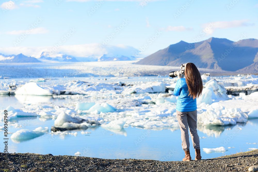 Photographer tourist woman taking photos with DSLR camera on travel on Iceland by Jokulsarlon glacial lagoon / glacier lake on Iceland. Happy tourist girl on travel in beautiful nature landscape. - obrazy, fototapety, plakaty 