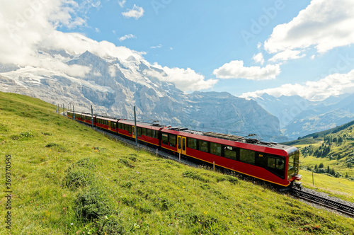 A cogwheel train travels on the railway from Jungfraujoch to Kleine Scheidegg on the green grassy hillside with Jungfrau & Monch mountains in background, in Bernese Highlands, Switzerland