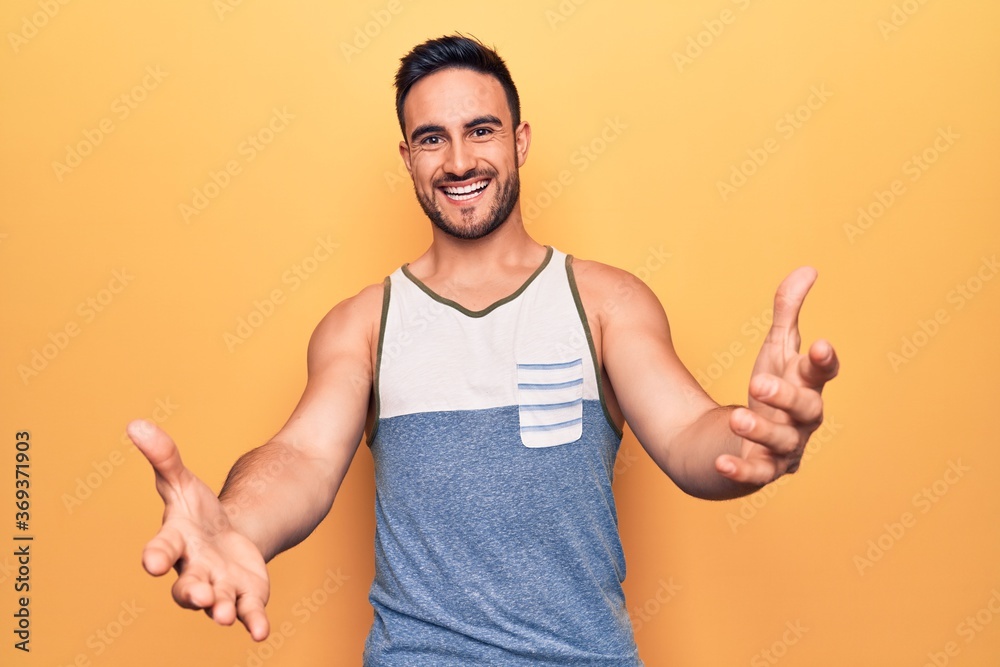 Young handsome man with beard wearing sleeveless t-shirt standing over yellow background looking at the camera smiling with open arms for hug. Cheerful expression embracing happiness. Young handsome man with beard wearing sleeveless t-shirt standing over yellow background looking at the camera smiling with open arms for hug. Cheerful expression embracing happiness.