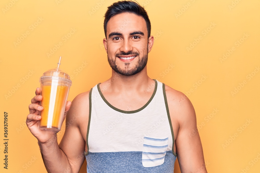 Handsome man with beard wearing sleeveless t-shirt drinking glass of healthy orange juice looking positive and happy standing and smiling with a confident smile showing teeth Handsome man with beard wearing sleeveless t-shirt drinking glass of healthy orange juice looking positive and happy standing and smiling with a confident smile showing teeth