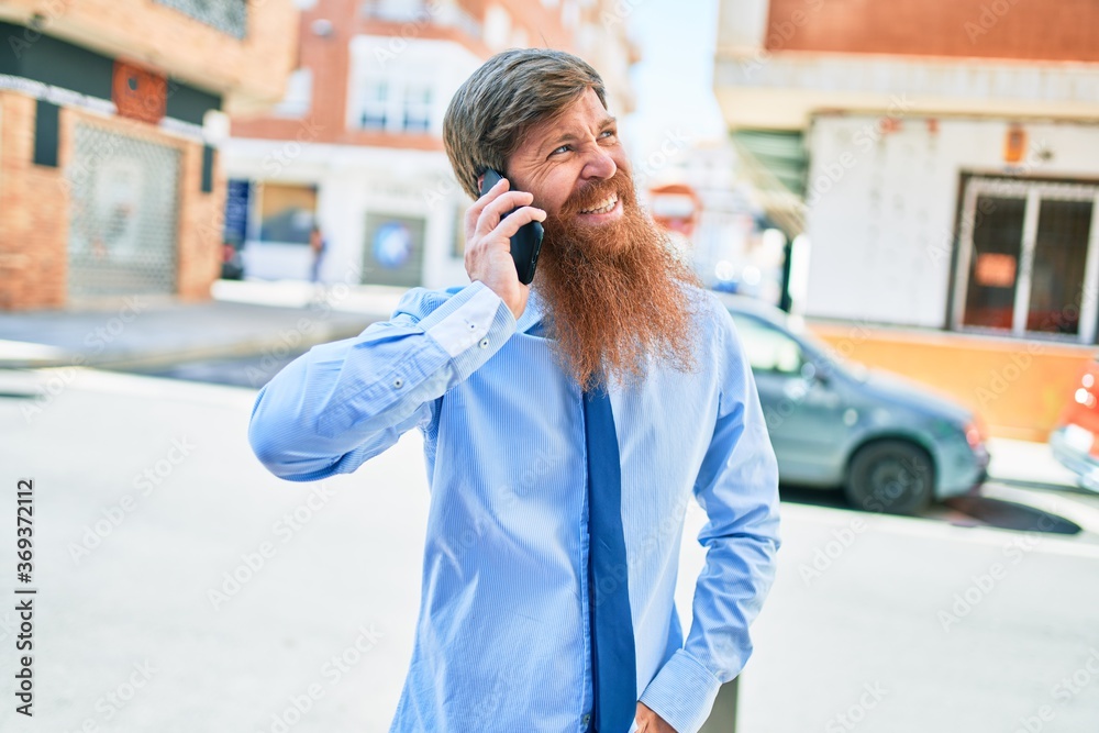 Young handsome redhead businessman smiling happy. Standing with smile on face having conversation talking on the smartphone at street of city.