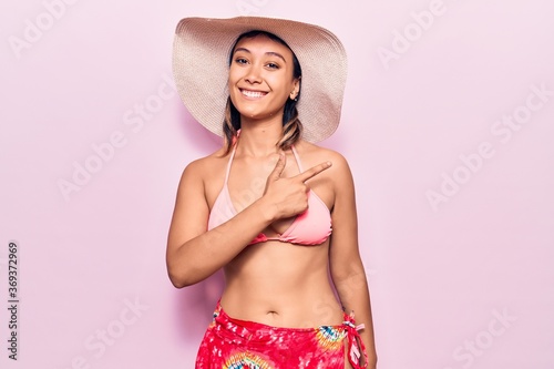 Young woman wearing bikini and hat cheerful with a smile of face pointing with hand and finger up to the side with happy and natural expression on face