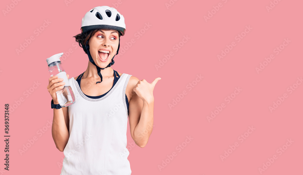 Beautiful young woman with short hair wearing bike helmet and holding water bottle pointing thumb up to the side smiling happy with open mouth Beautiful young woman with short hair wearing bike helmet and holding water bottle pointing thumb up to the side smiling happy with open mouth