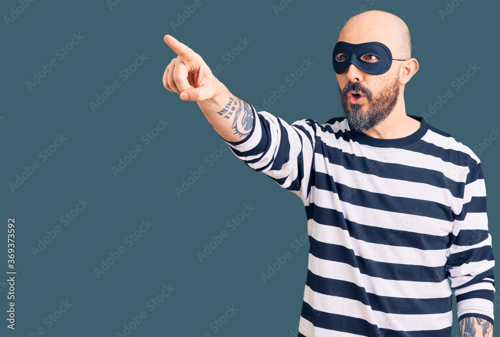 Naklejka premium Young handsome man wearing burglar mask pointing with finger surprised ahead, open mouth amazed expression, something on the front