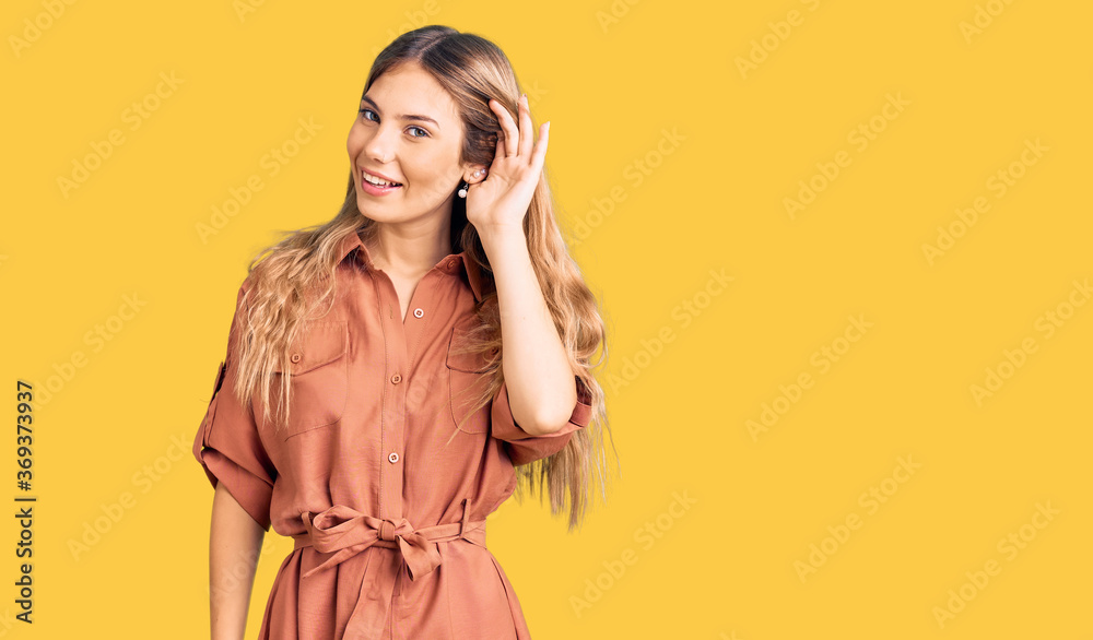 Beautiful caucasian woman with blonde hair wearing summer jumpsuit smiling with hand over ear listening an hearing to rumor or gossip. deafness concept. Beautiful caucasian woman with blonde hair wearing summer jumpsuit smiling with hand over ear listening an hearing to rumor or gossip. deafness concept.