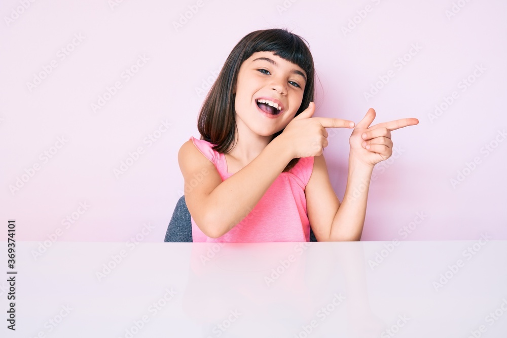Young little girl with bang wearing casual clothes sitting on the table smiling and looking at the camera pointing with two hands and fingers to the side. Young little girl with bang wearing casual clothes sitting on the table smiling and looking at the camera pointing with two hands and fingers to the side.