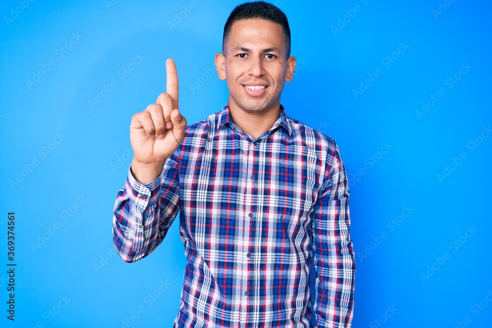 Young handsome latin man wearing casual clothes showing and pointing up with finger number one while smiling confident and happy. Young handsome latin man wearing casual clothes showing and pointing up with finger number one while smiling confident and happy.