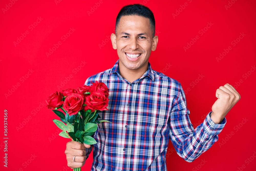 Young handsome latin man holding flowers screaming proud, celebrating victory and success very excited with raised arms Young handsome latin man holding flowers screaming proud, celebrating victory and success very excited with raised arms