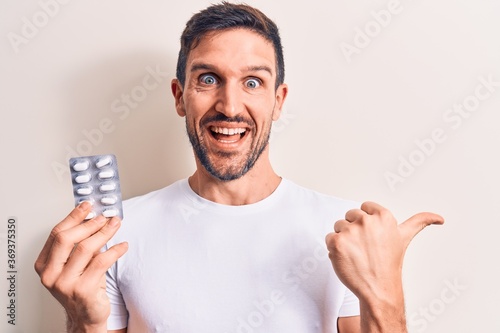 Young handsome man holding medicine pills standing over isolated white background pointing thumb up to the side smiling happy with open mouth