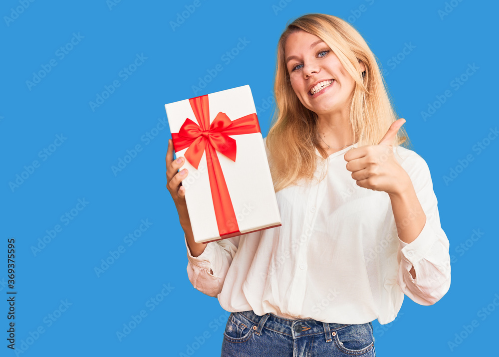 Young beautiful blonde woman holding gift smiling happy pointing with hand and finger to the side Young beautiful blonde woman holding gift smiling happy pointing with hand and finger to the side