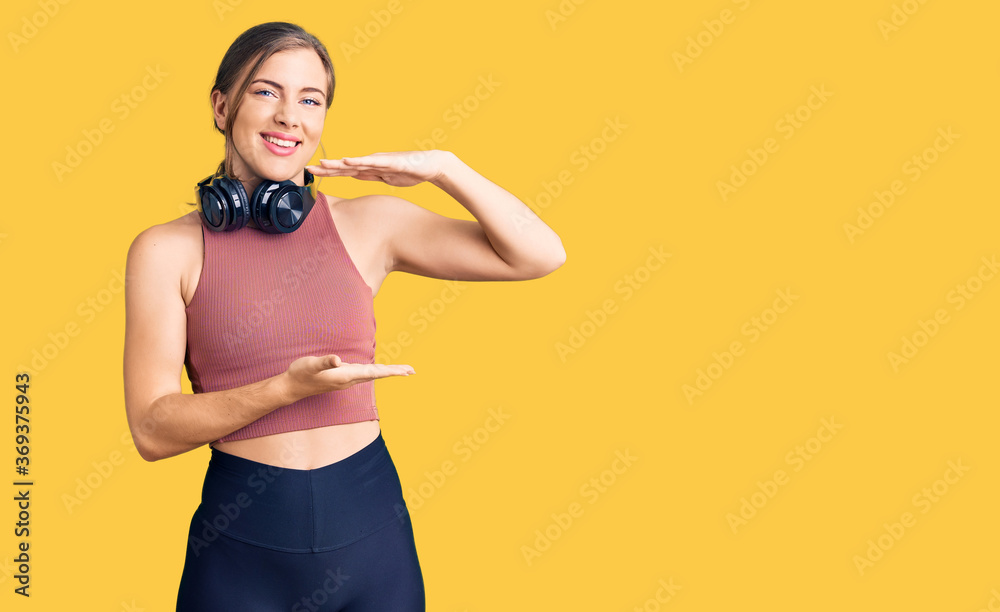 Beautiful caucasian young woman wearing gym clothes and using headphones gesturing with hands showing big and large size sign, measure symbol. smiling looking at the camera. measuring concept. Beautiful caucasian young woman wearing gym clothes and using headphones gesturing with hands showing big and large size sign, measure symbol. smiling looking at the camera. measuring concept.