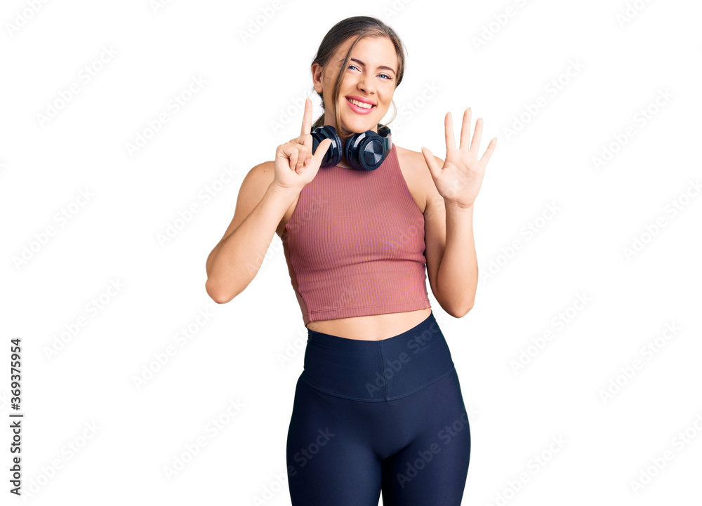 Beautiful caucasian young woman wearing gym clothes and using headphones showing and pointing up with fingers number seven while smiling confident and happy. Beautiful caucasian young woman wearing gym clothes and using headphones showing and pointing up with fingers number seven while smiling confident and happy.