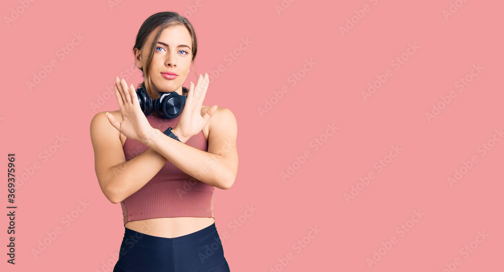 Obraz premium Beautiful caucasian young woman wearing gym clothes and using headphones rejection expression crossing arms and palms doing negative sign, angry face