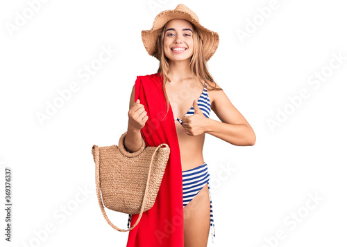 Young beautiful blonde woman wearing bikini and hat holding summer wicker handbag smiling happy and positive, thumb up doing excellent and approval sign photo