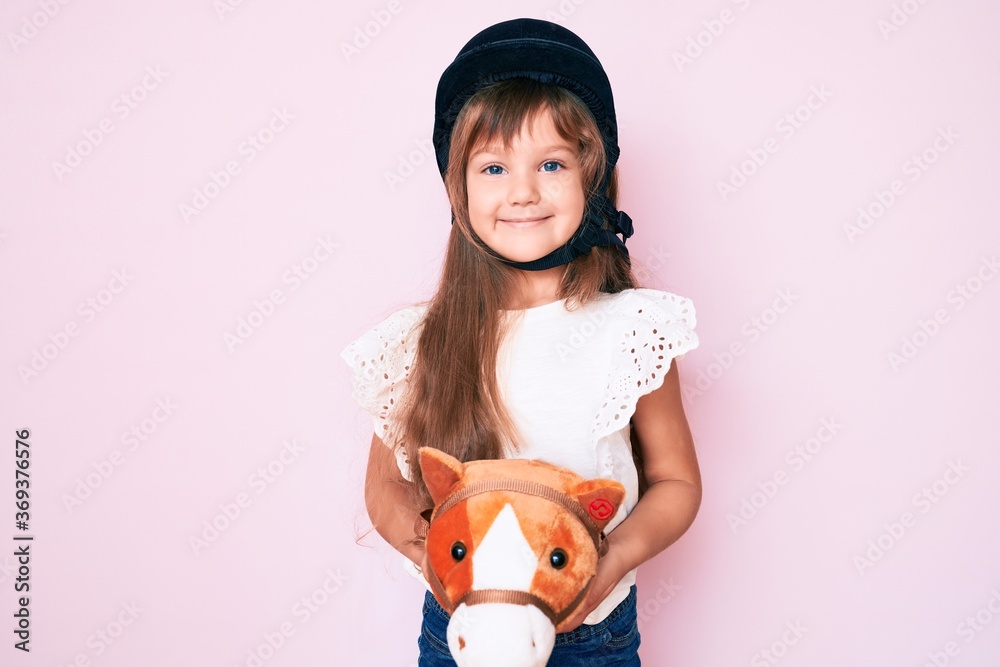 Little caucasian kid girl with long hair riding horse toy wearing vintage helmet looking positive and happy standing and smiling with a confident smile showing teeth