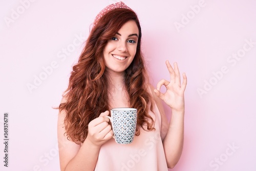 Young beautiful woman holding coffee doing ok sign with fingers, smiling friendly gesturing excellent symbol