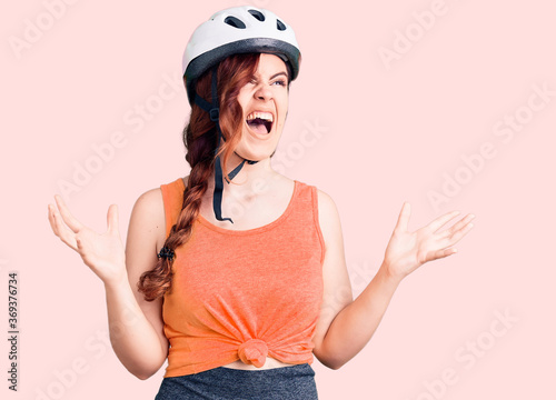 Young beautiful woman wearing bike helmet crazy and mad shouting and yelling with aggressive expression and arms raised. frustration concept.