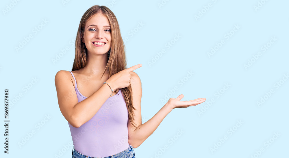 Young beautiful blonde woman wearing casual style with sleeveless shirt amazed and smiling to the camera while presenting with hand and pointing with finger. Young beautiful blonde woman wearing casual style with sleeveless shirt amazed and smiling to the camera while presenting with hand and pointing with finger.