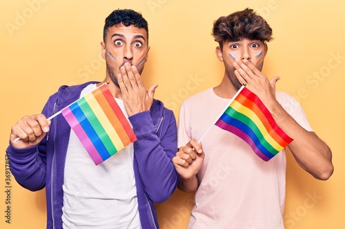 Young gay couple holding rainbow lgbtq flags covering mouth with hand, shocked and afraid for mistake. surprised expression