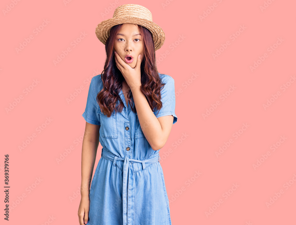 Young beautiful chinese girl wearing summer hat looking fascinated with disbelief, surprise and amazed expression with hands on chin Young beautiful chinese girl wearing summer hat looking fascinated with disbelief, surprise and amazed expression with hands on chin