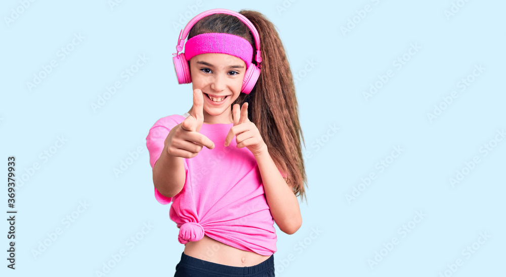 Cute hispanic child girl wearing gym clothes and using headphones pointing fingers to camera with happy and funny face. good energy and vibes. Cute hispanic child girl wearing gym clothes and using headphones pointing fingers to camera with happy and funny face. good energy and vibes.