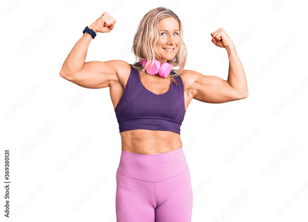 Middle age fit blonde woman wearing gym clothes and using headphones showing arms muscles smiling proud. fitness concept. Middle age fit blonde woman wearing gym clothes and using headphones showing arms muscles smiling proud. fitness concept.