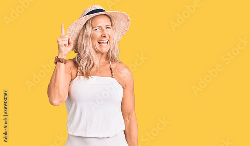 Middle age fit blonde woman wearing summer hat pointing finger up with successful idea. exited and happy. number one.