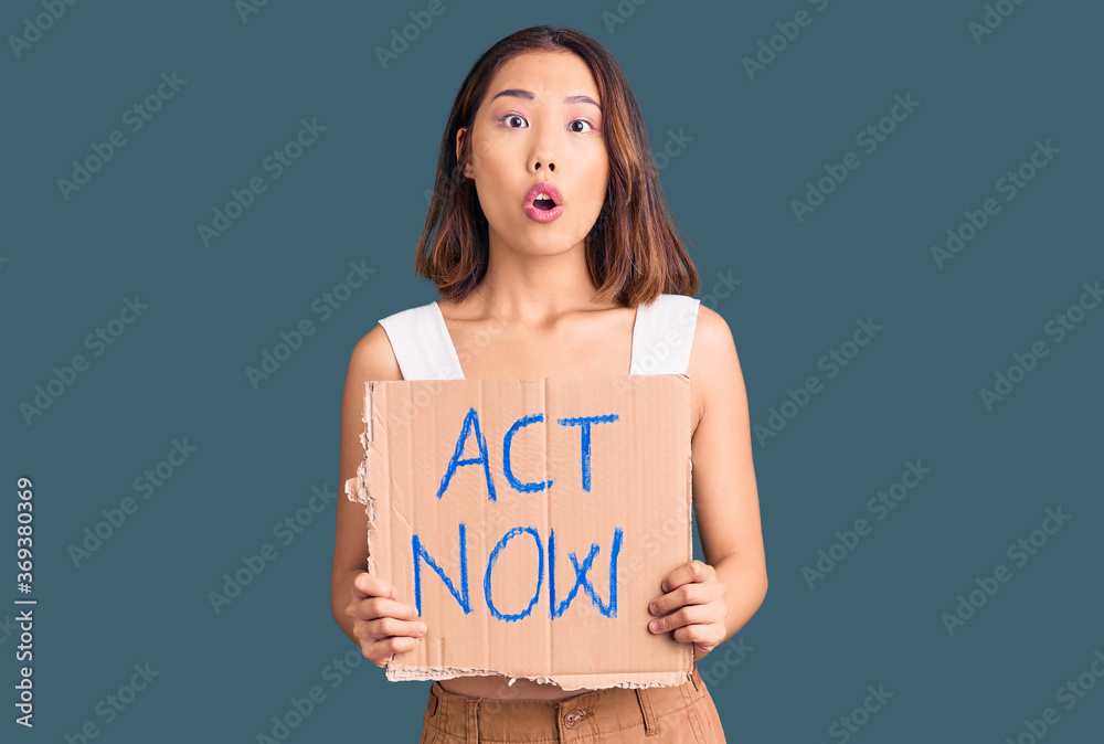 Young beautiful chinese girl holding act now banner scared and amazed with open mouth for surprise, disbelief face Young beautiful chinese girl holding act now banner scared and amazed with open mouth for surprise, disbelief face