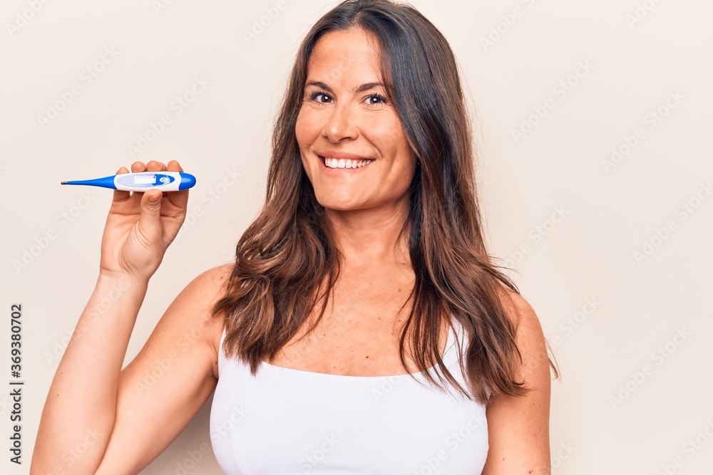 Beautiful brunette woman controlling temperature holding thermometer over white background looking positive and happy standing and smiling with a confident smile showing teeth