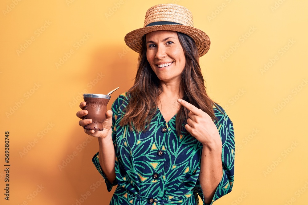 Young beautiful brunette woman wearing summer hat drinking cup of mate infusion beverage smiling happy pointing with hand and finger