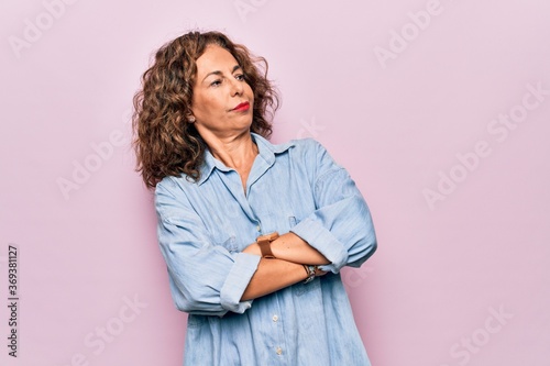 Middle age beautiful woman wearing casual denim shirt standing over pink background looking to the side with arms crossed convinced and confident