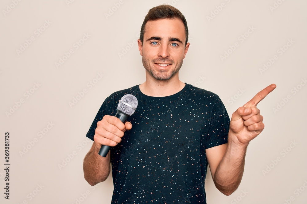 Young handsome singer man with blue eyes singing using microphone over white background very happy pointing with hand and finger to the side Young handsome singer man with blue eyes singing using microphone over white background very happy pointing with hand and finger to the side