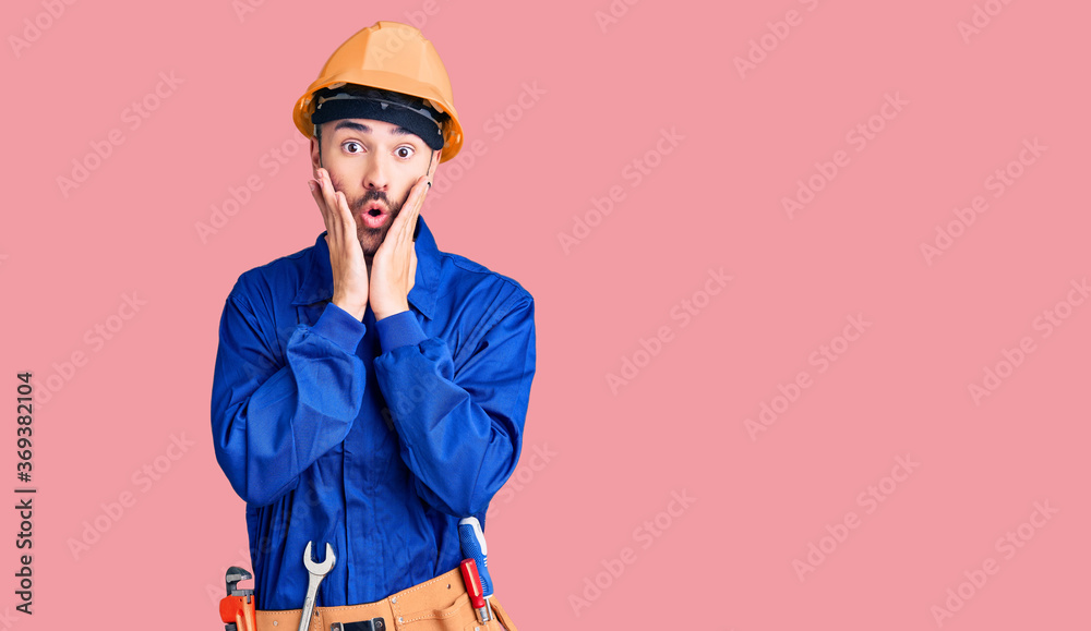 Young hispanic man wearing worker uniform afraid and shocked, surprise and amazed expression with hands on face