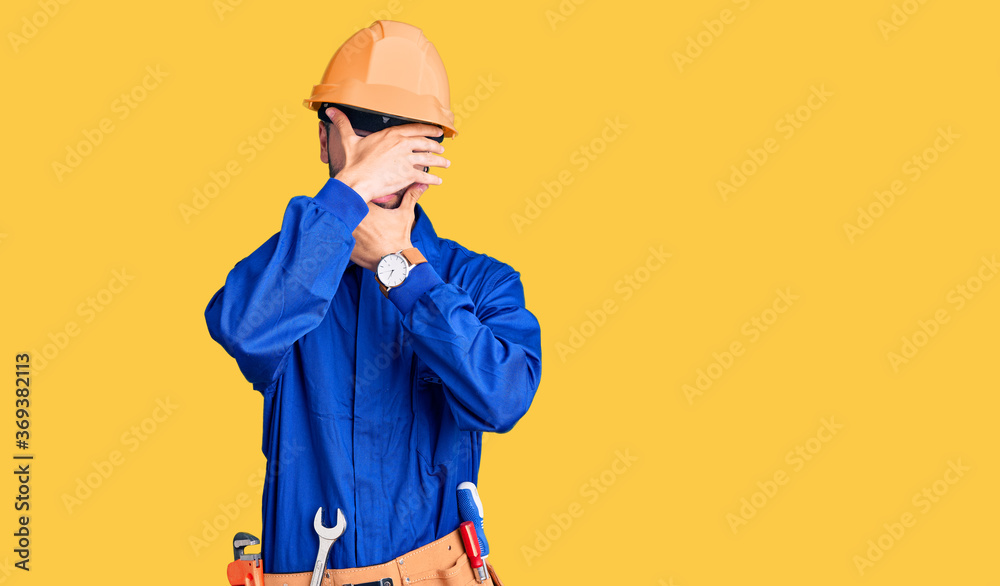 Young hispanic man wearing worker uniform covering eyes and mouth with hands, surprised and shocked. hiding emotion Young hispanic man wearing worker uniform covering eyes and mouth with hands, surprised and shocked. hiding emotion