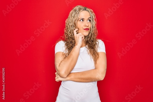 Young beautiful blonde woman wearing casual t-shirt standing over isolated red background with hand on chin thinking about question, pensive expression. Smiling with thoughtful face. Doubt concept.