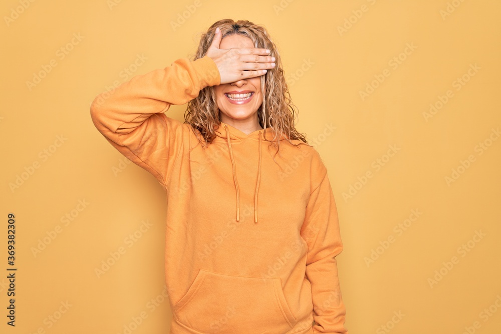 Young beautiful blonde sporty woman wearing casual sweatshirt over yellow background smiling and laughing with hand on face covering eyes for surprise. Blind concept. Young beautiful blonde sporty woman wearing casual sweatshirt over yellow background smiling and laughing with hand on face covering eyes for surprise. Blind concept.