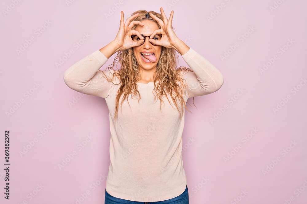 Young beautiful blonde woman wearing casual sweater and glasses over pink background doing ok gesture like binoculars sticking tongue out, eyes looking through fingers. Crazy expression. Young beautiful blonde woman wearing casual sweater and glasses over pink background doing ok gesture like binoculars sticking tongue out, eyes looking through fingers. Crazy expression.
