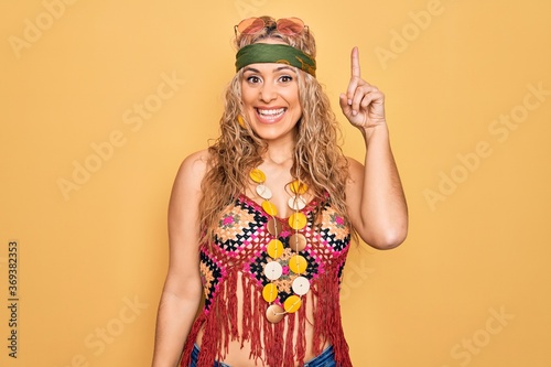 Beautiful blonde hippie woman wearing sunglasses and accessories over yellow background pointing finger up with successful idea. Exited and happy. Number one.
