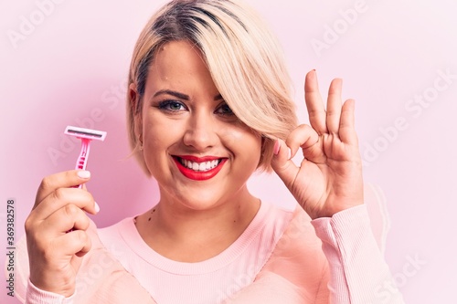 Young beautiful blonde plus size woman holding depilation razor over isolated pink background doing ok sign with fingers, smiling friendly gesturing excellent symbol