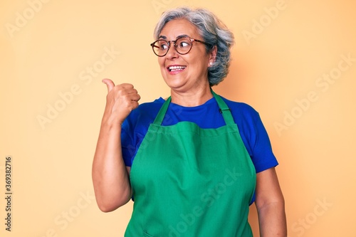 Senior hispanic woman wearing apron and glasses smiling with happy face looking and pointing to the side with thumb up.