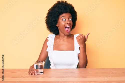 Young african american woman drinking glass of water pointing thumb up to the side smiling happy with open mouth