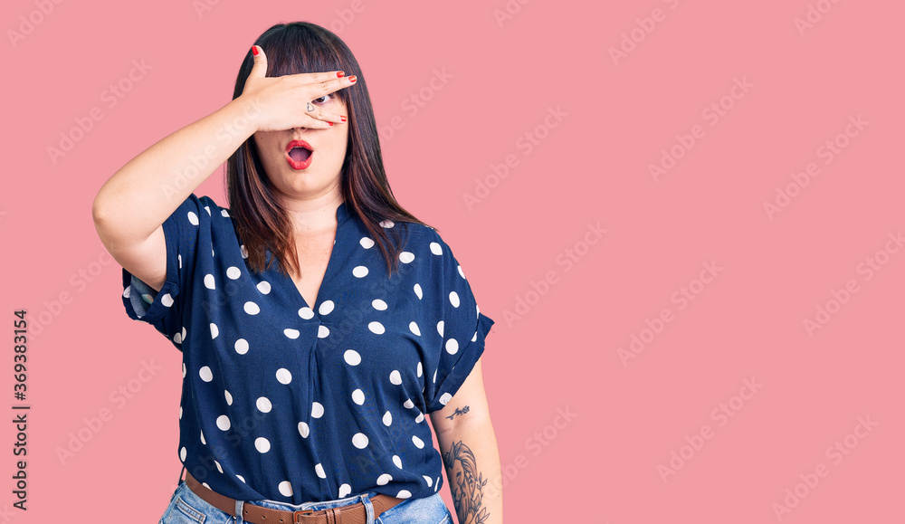 Young plus size woman wearing casual clothes peeking in shock covering face and eyes with hand, looking through fingers with embarrassed expression. Young plus size woman wearing casual clothes peeking in shock covering face and eyes with hand, looking through fingers with embarrassed expression.