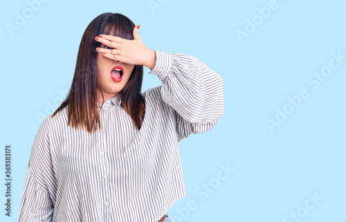 Young plus size woman wearing casual clothes peeking in shock covering face and eyes with hand, looking through fingers with embarrassed expression. © Krakenimages.com