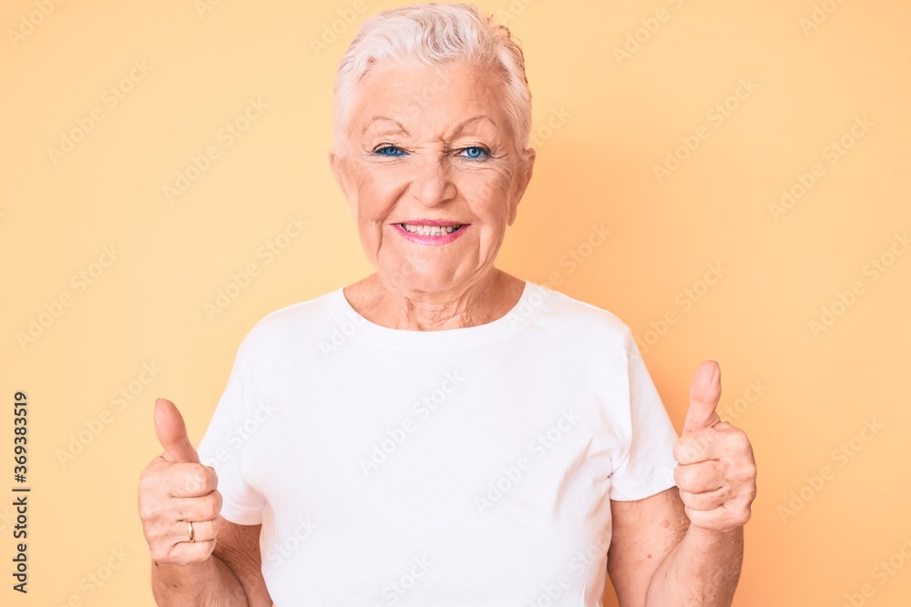 Senior beautiful woman with blue eyes and grey hair wearing classic white tshirt over yellow background success sign doing positive gesture with hand, thumbs up smiling and happy. cheerful expression. Senior beautiful woman with blue eyes and grey hair wearing classic white tshirt over yellow background success sign doing positive gesture with hand, thumbs up smiling and happy. cheerful expression.