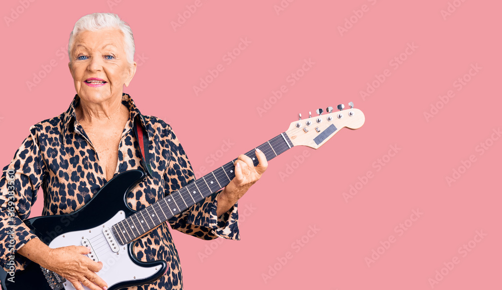 Senior beautiful woman with blue eyes and grey hair with modern look playing electric guitar looking positive and happy standing and smiling with a confident smile showing teeth