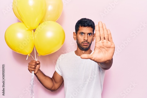 Young latin man holding balloons with open hand doing stop sign with serious and confident expression, defense gesture © Krakenimages.com