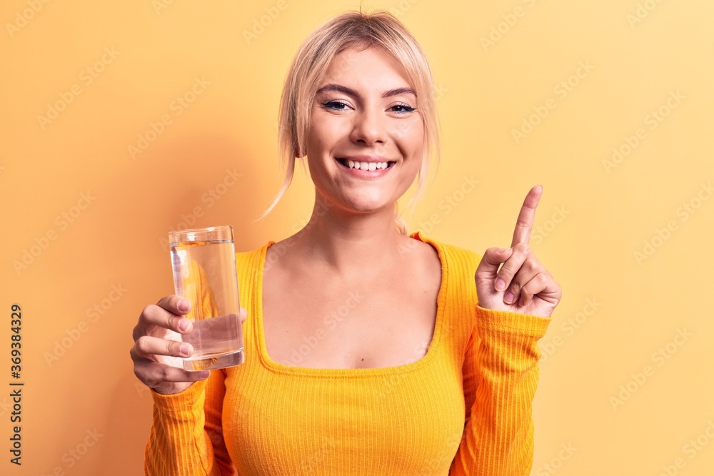 Young beautiful blonde woman drinking glass of water over isolated yellow background smiling with an idea or question pointing finger with happy face, number one Young beautiful blonde woman drinking glass of water over isolated yellow background smiling with an idea or question pointing finger with happy face, number one