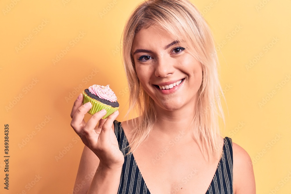 Young beautiful blonde woman eating sweet chocolate cupcake over isolated yellow background looking positive and happy standing and smiling with a confident smile showing teeth Young beautiful blonde woman eating sweet chocolate cupcake over isolated yellow background looking positive and happy standing and smiling with a confident smile showing teeth