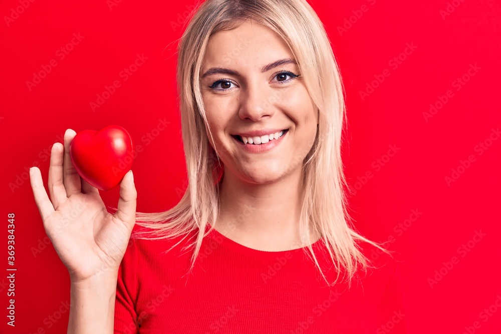 Young beautiful blonde woman holding plastic heart standing over isolated red background looking positive and happy standing and smiling with a confident smile showing teeth Young beautiful blonde woman holding plastic heart standing over isolated red background looking positive and happy standing and smiling with a confident smile showing teeth